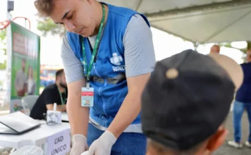 Central do Recomeço atende 100 pessoas no Pinheirinho Profissional de saúde da equipe do Consultório na Rua, vestindo colete azul, realiza atendimento e curativo no braço de um homem sentado durante a ação da Central do Recomeço no bairro Pinheirinho. Ao fundo, observa-se uma mesa de atendimento identificada com a placa do Cadastro Único.