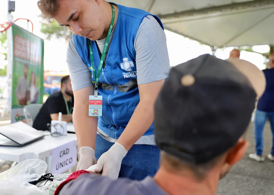 Profissional de saúde da equipe do Consultório na Rua, vestindo colete azul, realiza atendimento e curativo no braço de um homem sentado durante a ação da Central do Recomeço no bairro Pinheirinho. Ao fundo, observa-se uma mesa de atendimento identificada com a placa do Cadastro Único.
