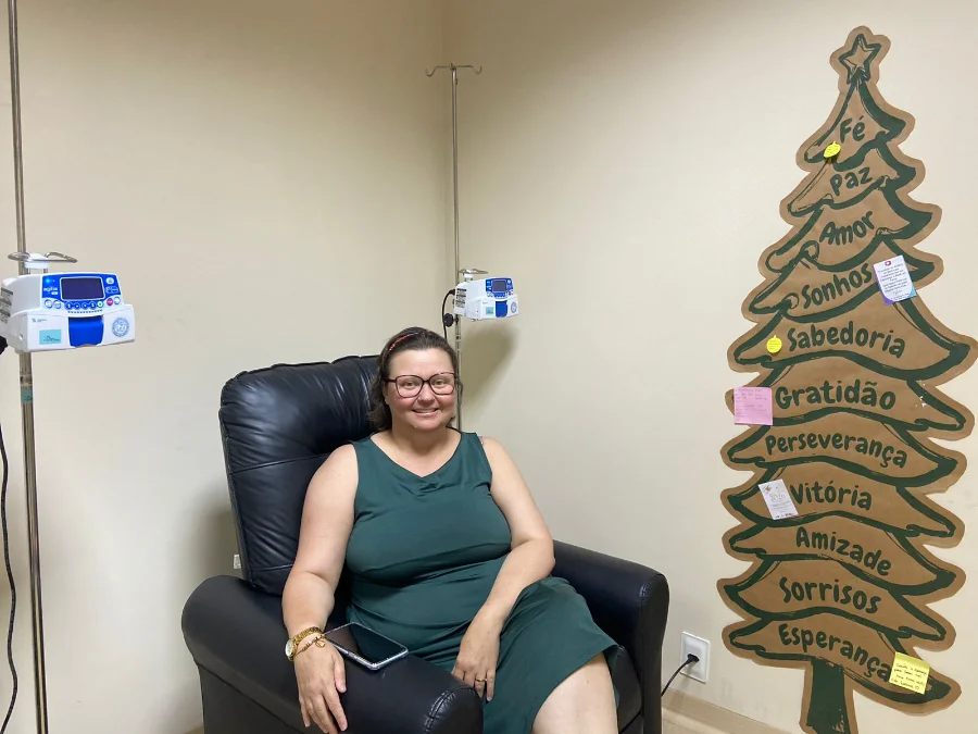 Paciente Andreia Moreira sorri sentada em poltrona de tratamento no CEPON ao lado de decoração com a palavra esperança, simbolizando o impacto positivo da pesquisa clínica contra o câncer em Santa Catarina.