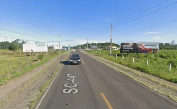 Ciclovia da SC-447: Empresa é habilitada para a obra Vista asfáltica da rodovia SC-447 em trecho plano, com vegetação e placas publicitárias nas margens. Sobre a pista, a inscrição "SC-447" identifica o local onde será implantada a futura ciclovia entre Araranguá e Arroio do Silva.