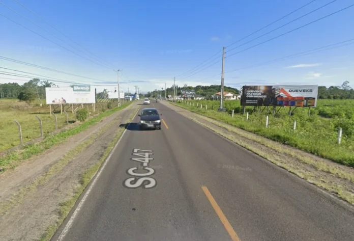 Ciclovia da SC-447 Empresa é habilitada para a obra Vista asfáltica da rodovia SC-447 em trecho plano, com vegetação e placas publicitárias nas margens. Sobre a pista, a inscrição "SC-447" identifica o local onde será implantada a futura ciclovia entre Araranguá e Arroio do Silva.