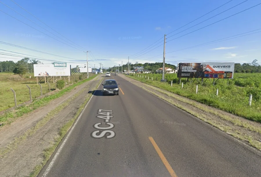 Ciclovia da SC-447 Empresa é habilitada para a obra Vista asfáltica da rodovia SC-447 em trecho plano, com vegetação e placas publicitárias nas margens. Sobre a pista, a inscrição