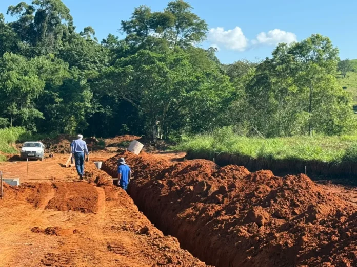 Cocal do Sul avança com esgoto para casas populares Fotografia de uma obra de infraestrutura em Cocal do Sul. Em um terreno de terra batida avermelhada, destaca-se uma vala profunda e extensa escavada para a instalação da rede de esgoto. Dois homens caminham pelo local da obra, e um carro branco está estacionado ao lado esquerdo. Ao fundo, uma densa área de mata verde sob um céu azul claro completa o cenário.