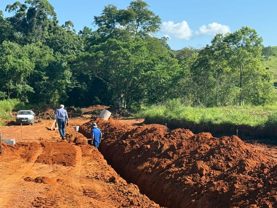 Fotografia de uma obra de infraestrutura em Cocal do Sul. Em um terreno de terra batida avermelhada, destaca-se uma vala profunda e extensa escavada para a instalação da rede de esgoto. Dois homens caminham pelo local da obra, e um carro branco está estacionado ao lado esquerdo. Ao fundo, uma densa área de mata verde sob um céu azul claro completa o cenário.