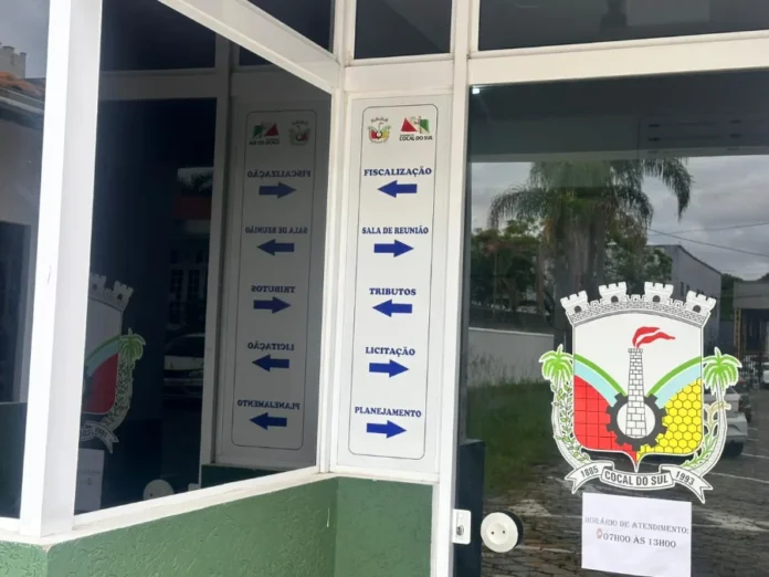 Cocal do Sul libera guias de taxas para empresas locais Fachada do setor administrativo da Prefeitura de Cocal do Sul, mostrando a porta de vidro com o brasão do município e placas indicativas para os departamentos de Tributos, Fiscalização e Planejamento.