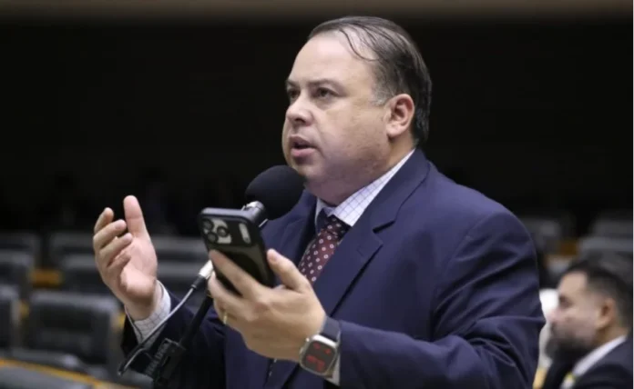 Comissão aprova incentivo à inovação em áreas com baixo IDH Foto de meio corpo de homem de pele clara, vestindo terno azul escuro e gravata vinho, falando ao microfone enquanto segura um celular na mão esquerda. Ele está em um ambiente legislativo, com fileiras de cadeiras desfocadas ao fundo.