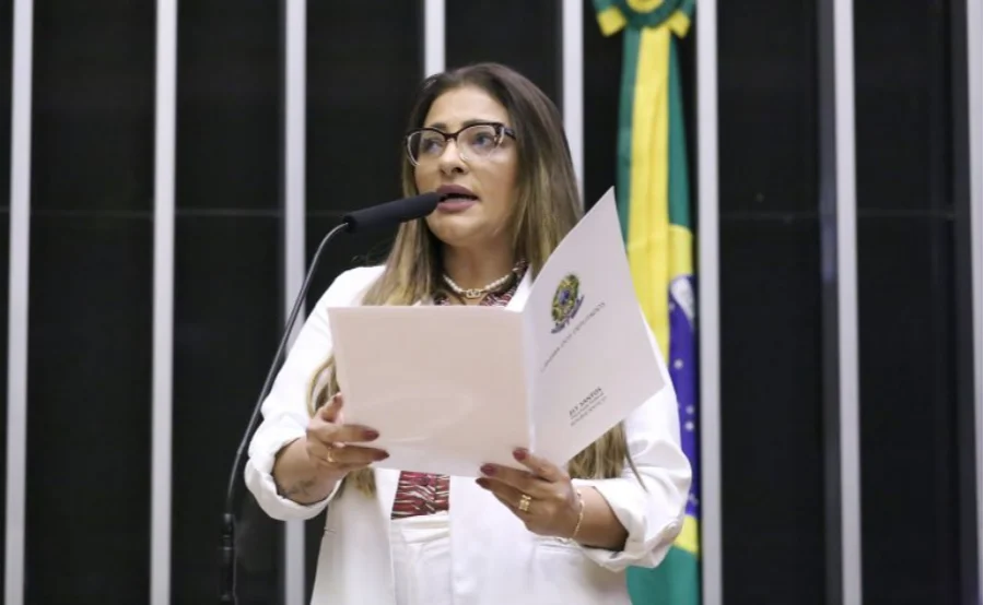 A deputada federal Ely Santos, relatora do projeto, discursa na Câmara dos Deputados segurando o parecer oficial. Ela é uma mulher de cabelos castanhos, usa óculos de grau e blazer branco. Ao fundo, vê-se parte da bandeira do Brasil. A imagem ilustra a aprovação da proposta que beneficia candidatas gestantes no processo da CNH.