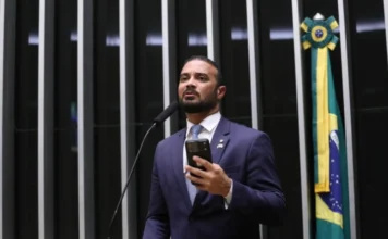 Comissão proíbe separar presos apenas por facção criminosa Deputado Capitão Alden, relator do projeto, discursa ao microfone na Câmara dos Deputados, vestindo terno azul e segurando um celular.