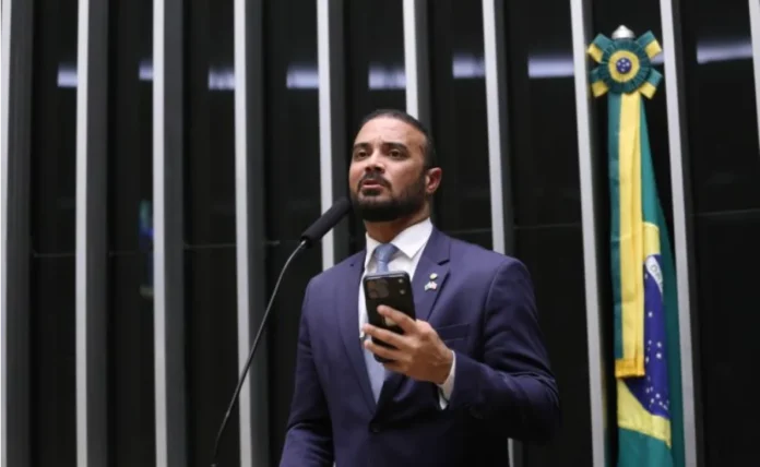 Comissão proíbe separar presos apenas por facção criminosa Deputado Capitão Alden, relator do projeto, discursa ao microfone na Câmara dos Deputados, vestindo terno azul e segurando um celular.
