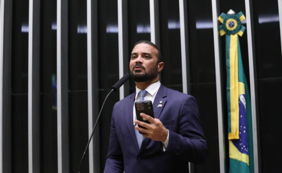 Deputado Capitão Alden, relator do projeto, discursa ao microfone na Câmara dos Deputados, vestindo terno azul e segurando um celular.