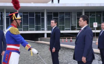Congresso abre 4ª sessão legislativa nesta segunda-feira A imagem registra um momento do rito militar na rampa do Congresso Nacional. À esquerda, em primeiro plano, um integrante da unidade Dragões da Independência, vestindo uniforme histórico azul com detalhes em vermelho e dourado, presta continência segurando uma espada. À direita, o presidente da Câmara dos Deputados, Hugo Motta, e o presidente do Senado, Davi Alcolumbre, vestem ternos escuros e observam formalmente a cerimônia. Ao fundo, é possível ver a arquitetura de vidro do Palácio do Congresso e o espelho d'água.