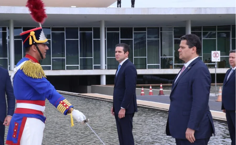 A imagem registra um momento do rito militar na rampa do Congresso Nacional. À esquerda, em primeiro plano, um integrante da unidade Dragões da Independência, vestindo uniforme histórico azul com detalhes em vermelho e dourado, presta continência segurando uma espada. À direita, o presidente da Câmara dos Deputados, Hugo Motta, e o presidente do Senado, Davi Alcolumbre, vestem ternos escuros e observam formalmente a cerimônia. Ao fundo, é possível ver a arquitetura de vidro do Palácio do Congresso e o espelho d'água.