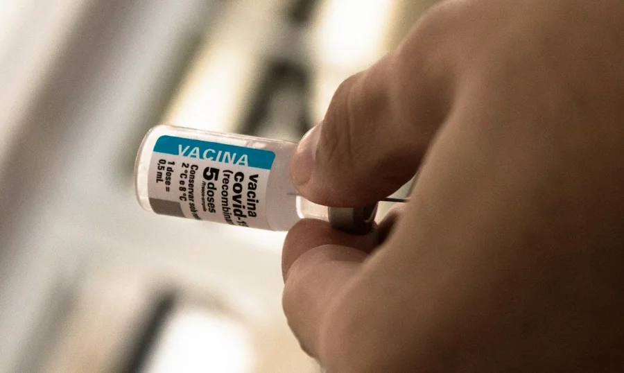 Covid-19 5 anos de vacina e os desafios da baixa adesão Close-up em tons quentes de uma mão segurando um frasco de vacina contra a covid-19 (recombinante). O rótulo em destaque mostra as informações '5 doses' e orientações de armazenamento entre 2°C e 8°C. A imagem simboliza a disponibilidade do imunizante e a continuidade da campanha de vacinação no Brasil cinco anos após seu início.