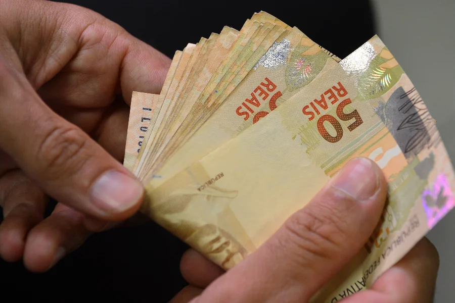 Crédito em SC prioriza capital de giro e acende alerta Mãos seguram um maço de notas de 50 reais, ilustrando recursos financeiros e o uso do dinheiro no contexto econômico de crédito e endividamento abordado na matéria.