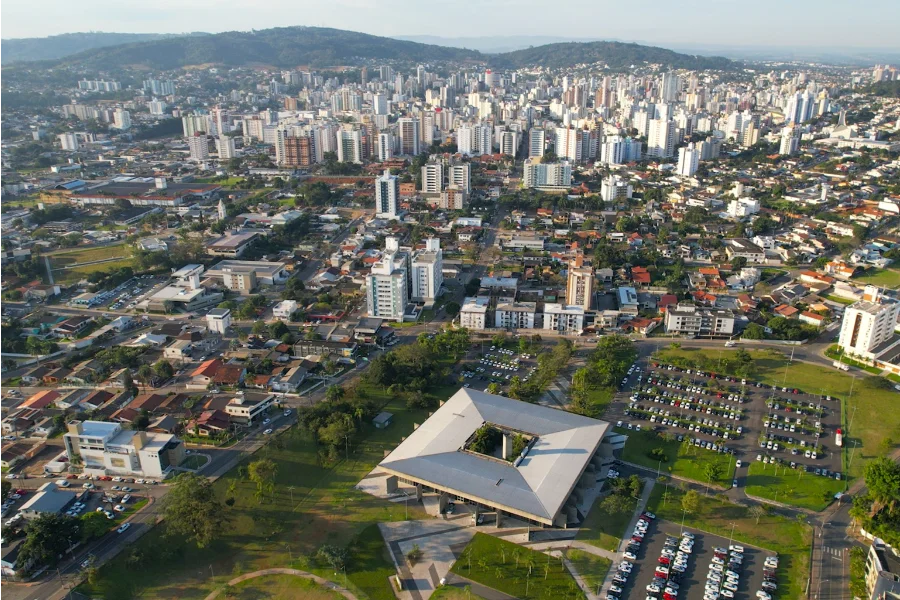 Criciúma 146 anos Veja o que abre e fecha no feriado Vista aérea da cidade de Criciúma em um dia ensolarado. No centro, destaca-se o Paço Municipal, um edifício quadrado com pátio interno cercado por estacionamentos e áreas verdes; ao fundo, o horizonte exibe a densa verticalização urbana com diversos prédios.