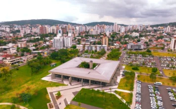 Criciúma abre concurso na saúde: salários até R$ 16,6 mil Vista aérea do Paço Municipal de Criciúma, sede da prefeitura, destacando a arquitetura quadrada e moderna do prédio administrativo em primeiro plano, cercado por jardins e um amplo estacionamento. Ao fundo, observa-se o horizonte urbano da cidade com diversos edifícios sob um céu nublado. A imagem representa o centro administrativo responsável pelo lançamento do novo concurso público da saúde.