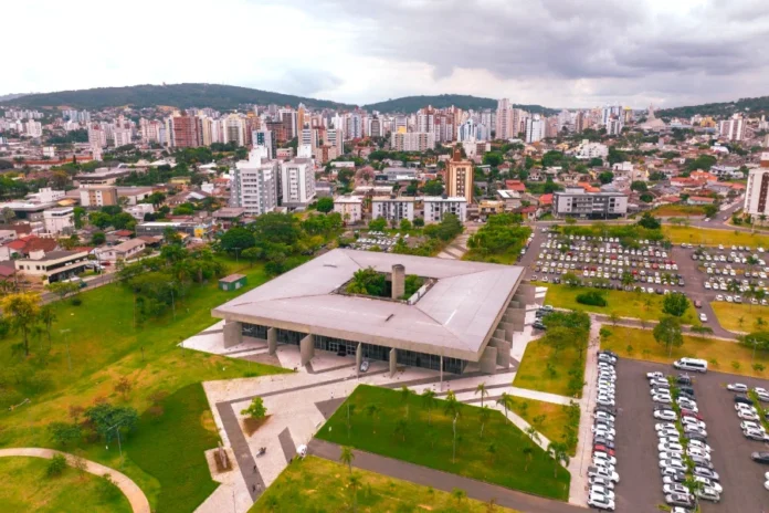 Criciúma abre concurso na saúde salários até R$ 16,6 mil Vista aérea do Paço Municipal de Criciúma, sede da prefeitura, destacando a arquitetura quadrada e moderna do prédio administrativo em primeiro plano, cercado por jardins e um amplo estacionamento. Ao fundo, observa-se o horizonte urbano da cidade com diversos edifícios sob um céu nublado. A imagem representa o centro administrativo responsável pelo lançamento do novo concurso público da saúde.