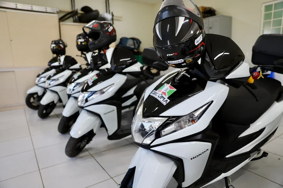 Fileira de novas motocicletas brancas (tipo scooter) da Prefeitura de Criciúma, estacionadas em uma garagem interna com capacetes pretos sobre os assentos. A moto em primeiro plano destaca o adesivo com o brasão do município. A nova frota foi adquirida para agilizar o trabalho dos Agentes de Combate às Endemias contra a dengue.