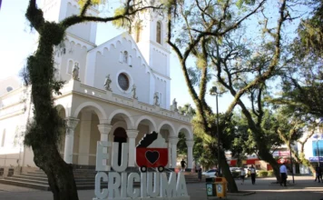 Criciúma celebra 146 anos de colonização nesta terça Vista frontal da Catedral São José em Criciúma, com árvores altas em primeiro plano e o letreiro "EU ❤️ CRICIÚMA" instalado na praça em frente à igreja.