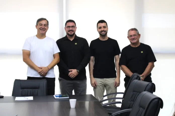 Criciúma empossa novos gestores de Obras e Comunicação Quatro homens em pé, lado a lado, sorrindo em uma sala de reuniões. O prefeito Vagner Espindola (à esquerda, de branco) e o vice Salésio Lima (à direita, de preto) ladeiam os novos gestores João Paulo Casagrande e Renan Medeiros. À frente deles, uma mesa de reuniões preta com cadeiras de escritório.