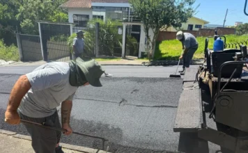 Criciúma fecha 2025 com 167 ruas pavimentadas e 65km de obras Três operários com uniformes cinzas trabalham na pavimentação asfáltica de uma rua; um deles, em primeiro plano, usa chapéu de aba larga e espalha a massa asfáltica com um rodo de metal ao lado de uma máquina pavimentadora.