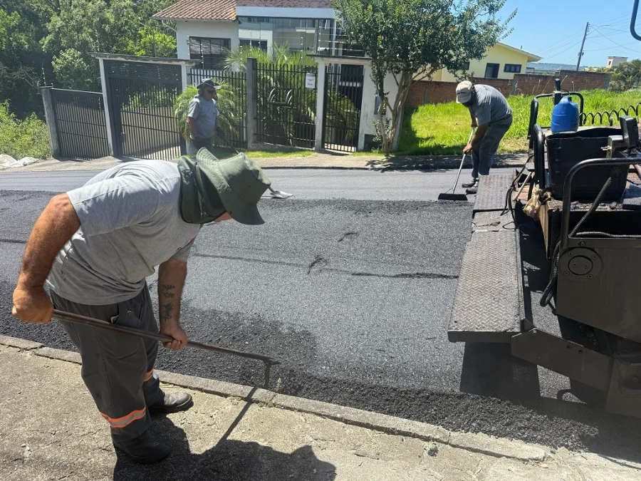 Criciúma fecha 2025 com 167 ruas pavimentadas e 65km de obras Três operários com uniformes cinzas trabalham na pavimentação asfáltica de uma rua; um deles, em primeiro plano, usa chapéu de aba larga e espalha a massa asfáltica com um rodo de metal ao lado de uma máquina pavimentadora.