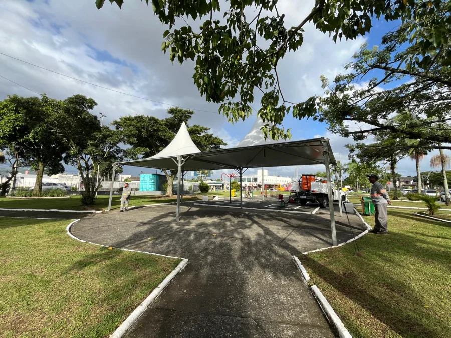 Estrutura de tendas brancas sendo montada na praça do bairro Pinheirinho, em Criciúma, para a ação social da prefeitura que oferecerá serviços de saúde, assistência social e emprego nos dias 20 e 21. Trabalhadores e um caminhão estão no local.