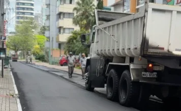 Criciúma inicia força-tarefa para obras em vias centrais Caminhão caçamba e equipe de trabalhadores realizam obra de pavimentação asfáltica em rua urbana, ilustrando a força-tarefa de revitalização de vias da Prefeitura de Criciúma.