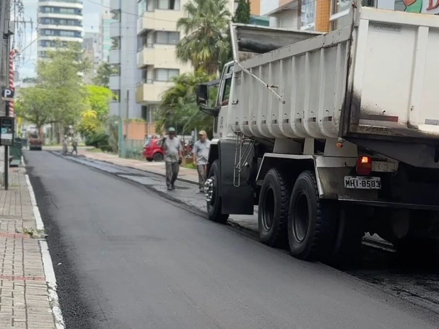 Criciúma inicia força-tarefa para obras em vias centrais Caminhão caçamba e equipe de trabalhadores realizam obra de pavimentação asfáltica em rua urbana, ilustrando a força-tarefa de revitalização de vias da Prefeitura de Criciúma.