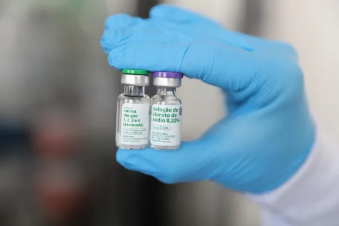 Criciúma inicia vacinação contra dengue para jovens na segunda Fotografia em close-up mostra uma mão utilizando luva de procedimento azul e segurando dois pequenos frascos de vidro. O frasco à esquerda possui rótulo verde e branco com a inscrição "vacina dengue 1, 2, 3 e 4 (atenuada)", e o frasco à direita possui rótulo roxo e branco identificando a "solução de cloreto de sódio 0,22%". A imagem, com fundo desfocado, ilustra o imunizante que será utilizado na campanha de vacinação em Criciúma.