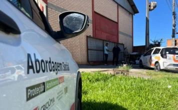 Criciúma interna sete dependentes químicos em um único dia Fotografia em ângulo lateral de um veículo oficial da Prefeitura de Criciúma. Em destaque, no primeiro plano, está o adesivo na lateral do carro com os dizeres "Abordagem Social" e as palavras "Proteção, Acolhimento e Inclusão", acompanhados pelo brasão do município. Ao fundo, em uma área externa com gramado e um edifício de tijolos, é possível ver integrantes da equipe técnica realizando um atendimento de rua próximo a outro veículo da administração municipal.