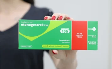 Criciúma oferta implante contraceptivo gratuito na rede Em plano detalhe, mãos femininas seguram a embalagem do implante contraceptivo subdérmico Implanon NXT. A caixa, nas cores verde e vermelho, destaca o princípio ativo etonogestrel 68 mg e traz os logotipos do SUS, do Ministério da Saúde e do Governo Federal, ilustrando a distribuição gratuita do método na rede pública.