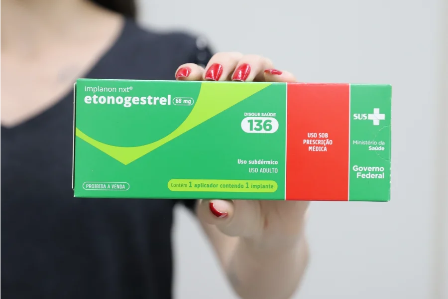 Criciúma oferta implante contraceptivo gratuito na rede Em plano detalhe, mãos femininas seguram a embalagem do implante contraceptivo subdérmico Implanon NXT. A caixa, nas cores verde e vermelho, destaca o princípio ativo etonogestrel 68 mg e traz os logotipos do SUS, do Ministério da Saúde e do Governo Federal, ilustrando a distribuição gratuita do método na rede pública.