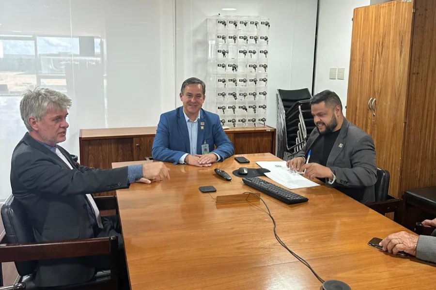 Foto de uma reunião em um escritório em Brasília, onde três homens estão sentados ao redor de uma grande mesa de madeira. O prefeito Vagner Espindola está entre eles, discutindo a captação de recursos para a obra do canal auxiliar de Criciúma.