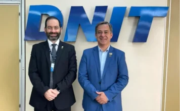 Criciúma propõe elevar ferrovia em sete metros ao DNIT Fotografia registra o prefeito de Criciúma, Vagner Espindola (à direita, em terno azul), ao lado do diretor de Infraestrutura Ferroviária do DNIT, Eloi Angelo Palma Filho (à esquerda, em terno preto). Eles estão em pé, lado a lado, à frente de uma parede clara que exibe o logotipo metálico azul do DNIT. A imagem captura o encontro oficial em Brasília onde foi discutido o projeto de elevação da linha férrea no perímetro urbano de Criciúma.