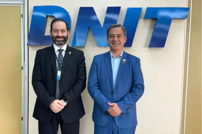 Criciúma propõe elevar ferrovia em 7 metros ao DNIT Fotografia registra o prefeito de Criciúma, Vagner Espindola (à direita, em terno azul), ao lado do diretor de Infraestrutura Ferroviária do DNIT, Eloi Angelo Palma Filho (à esquerda, em terno preto). Eles estão em pé, lado a lado, à frente de uma parede clara que exibe o logotipo metálico azul do DNIT. A imagem captura o encontro oficial em Brasília onde foi discutido o projeto de elevação da linha férrea no perímetro urbano de Criciúma.
