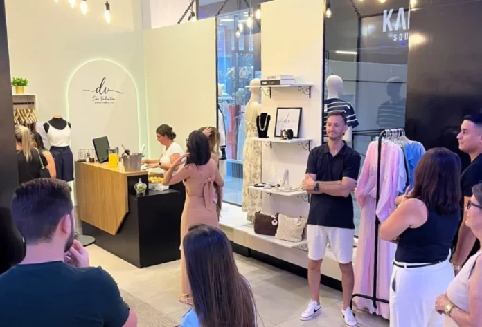 Dai Valentim inaugura loja no Sombrio Moda Shopping Interior de uma loja de roupas femininas durante um evento, com mulheres circulando, uma mesa redonda branca com doces e salgados, e um balcão de atendimento com bebidas.