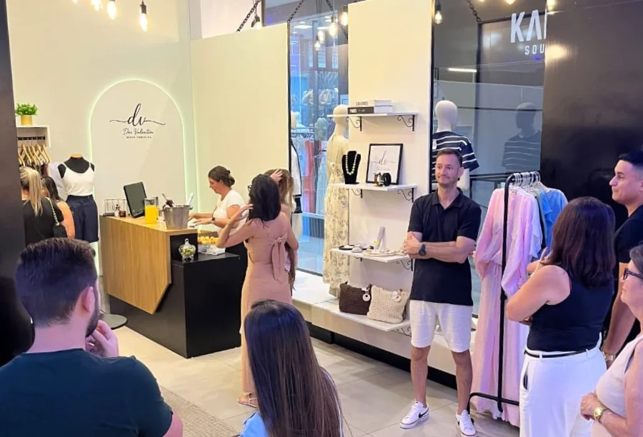 Dai Valentim inaugura loja no Sombrio Moda Shopping Interior de uma loja de roupas femininas durante um evento, com mulheres circulando, uma mesa redonda branca com doces e salgados, e um balcão de atendimento com bebidas.