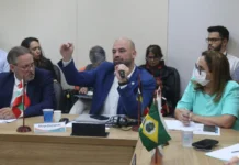 Diogo Demarchi debate saúde digital em assembleia no Rio Fotografia em plano médio do secretário Diogo Demarchi durante a assembleia do Conass. Ele é um homem calvo, com barba, veste terno azul e camisa branca. Ele segura um microfone com a mão esquerda e gesticula com a mão direita fechada, demonstrando ênfase em sua fala. À sua frente, sobre uma mesa de madeira, há uma placa de identificação com seu nome e as bandeiras de Santa Catarina e do Brasil. Outros participantes da reunião aparecem ao fundo e nas laterais da mesa.