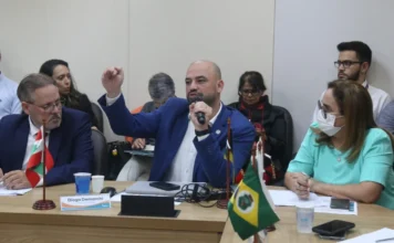 Diogo Demarchi debate saúde digital em assembleia no Rio Fotografia em plano médio do secretário Diogo Demarchi durante a assembleia do Conass. Ele é um homem calvo, com barba, veste terno azul e camisa branca. Ele segura um microfone com a mão esquerda e gesticula com a mão direita fechada, demonstrando ênfase em sua fala. À sua frente, sobre uma mesa de madeira, há uma placa de identificação com seu nome e as bandeiras de Santa Catarina e do Brasil. Outros participantes da reunião aparecem ao fundo e nas laterais da mesa.