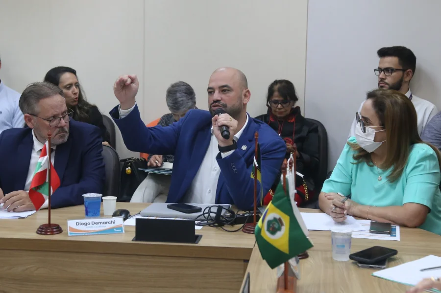 Fotografia em plano médio do secretário Diogo Demarchi durante a assembleia do Conass. Ele é um homem calvo, com barba, veste terno azul e camisa branca. Ele segura um microfone com a mão esquerda e gesticula com a mão direita fechada, demonstrando ênfase em sua fala. À sua frente, sobre uma mesa de madeira, há uma placa de identificação com seu nome e as bandeiras de Santa Catarina e do Brasil. Outros participantes da reunião aparecem ao fundo e nas laterais da mesa.