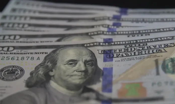 Dólar cai para R$ 5,40 e Bolsa sobe com foco na Venezuela Fotografia em close-up de várias cédulas de 100 dólares americanos espalhadas. A imagem foca no rosto de Benjamin Franklin em uma das notas em primeiro plano, com outras notas levemente desfocadas ao fundo. Ilustra o recuo da moeda para R$ 5,40.