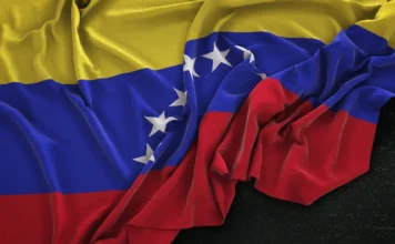 EUA apreendem petroleiros em rota para a Venezuela Close-up da bandeira da Venezuela com tecido drapeado sobre fundo preto. A imagem ilustra as tensões diplomáticas e o bloqueio econômico ao petróleo venezuelano citado na reportagem.