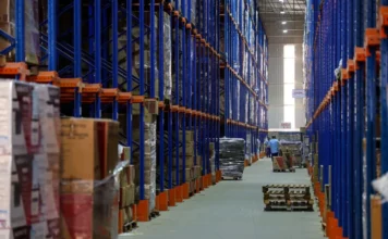 Economia de SC cresce 4,9% e lidera ranking nacional Corredor de um grande centro de distribuição logístico com prateleiras altas abastecidas de mercadorias e trabalhadores ao fundo, ilustrando o crescimento dos setores de comércio e indústria em Santa Catarina.