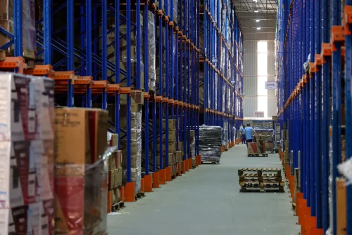 Economia de SC cresce 4,9% e lidera ranking nacional Corredor de um grande centro de distribuição logístico com prateleiras altas abastecidas de mercadorias e trabalhadores ao fundo, ilustrando o crescimento dos setores de comércio e indústria em Santa Catarina.