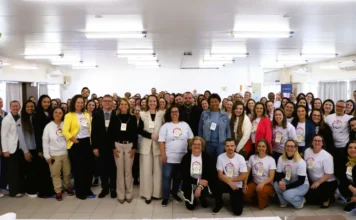 Educação em saúde atinge 45 mil profissionais em SC Grande grupo de dezenas de adultos, majoritariamente mulheres, posando sorridentes para uma foto coletiva em um auditório bem iluminado. Muitos usam camisetas brancas com logotipos coloridos e crachás, representando os profissionais de saúde, residentes, alunos e a equipe técnica da Escola de Saúde Pública de Santa Catarina (ESPSC) mencionados no balanço de atividades de 2025.