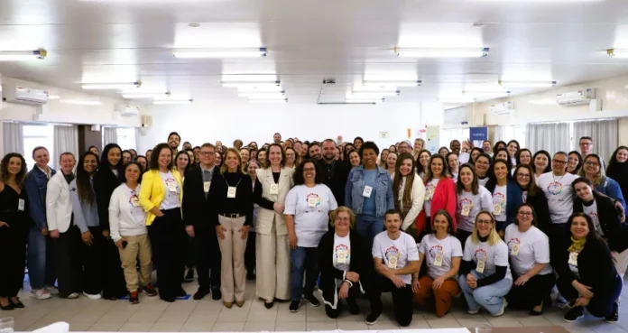Educação em saúde atinge 45 mil profissionais em SC Grande grupo de dezenas de adultos, majoritariamente mulheres, posando sorridentes para uma foto coletiva em um auditório bem iluminado. Muitos usam camisetas brancas com logotipos coloridos e crachás, representando os profissionais de saúde, residentes, alunos e a equipe técnica da Escola de Saúde Pública de Santa Catarina (ESPSC) mencionados no balanço de atividades de 2025.