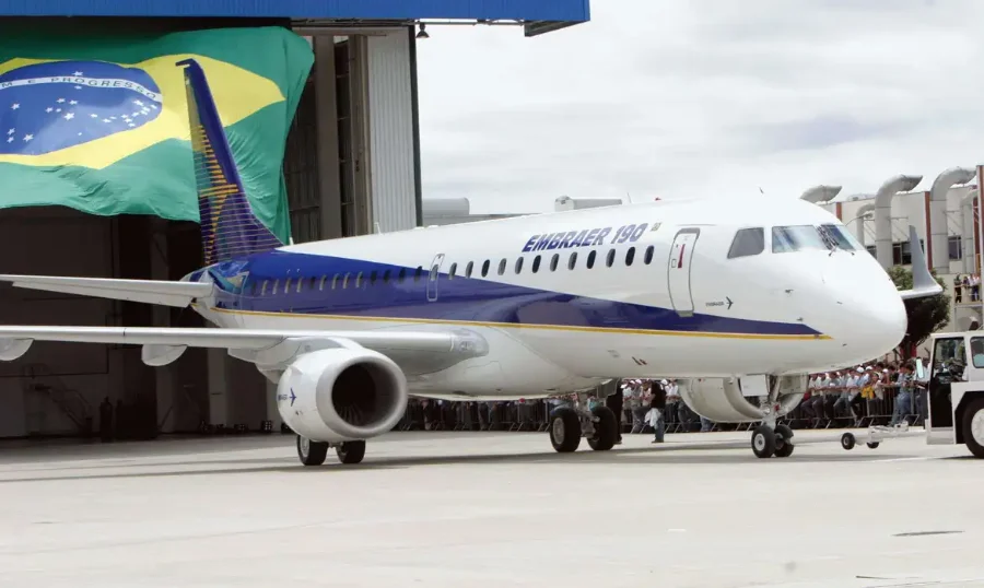 Embraer bate recorde com US$ 31,6 bilhões em pedidos Aeronave Embraer 190, nas cores branca e azul, posicionada no pátio em frente a um hangar onde se vê uma grande bandeira do Brasil ao fundo. Diversas pessoas acompanham a movimentação do jato, simbolizando o sucesso e a produção tecnológica da fabricante brasileira.