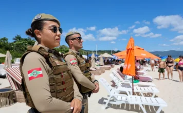 Estação Verão: indicadores de crimes caem em SC Policiais militares de Santa Catarina, um homem e uma mulher, realizam patrulhamento na areia da praia durante a Operação Estação Verão, com banhistas e guarda-sóis ao fundo sob céu azul.