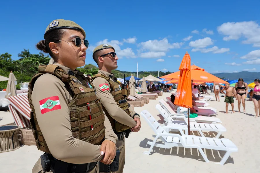 Policiais militares de Santa Catarina, um homem e uma mulher, realizam patrulhamento na areia da praia durante a Operação Estação Verão, com banhistas e guarda-sóis ao fundo sob céu azul.
